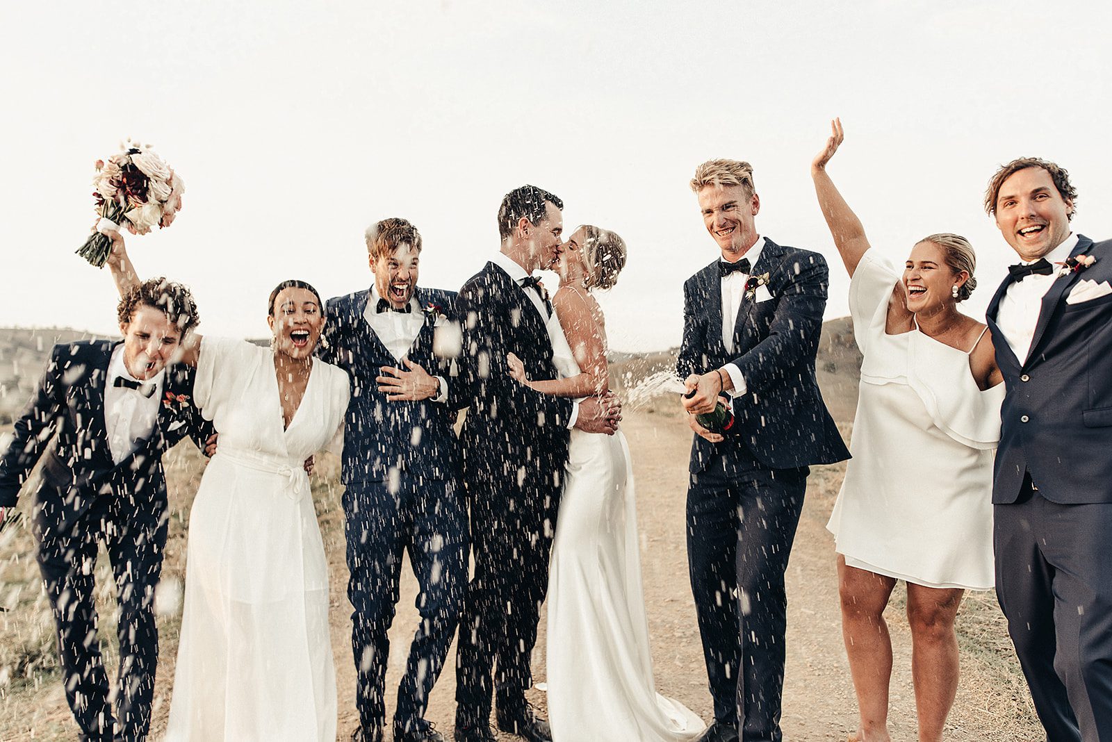 A bridal party in full celebration mode. Champagne is flowing in the air as the bride and groom Kiss passionately, their bridal party is having fun in this scene.