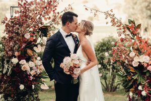 A beautiful wedding arbour with a bride and groom kissing, she is holding a wedding bouquet in a long slim wedding dress.