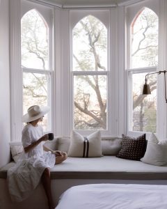 woman looking out the bay windows with a cup of tea in linen skirt on bay window seat with comfy cushions