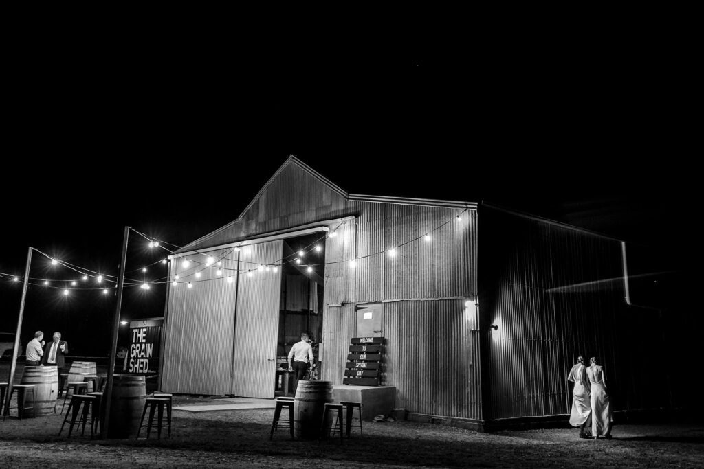 Stunning image od two brides heading around the back of the shed and I'm hoping it's the fathers of the bride talking at the other side of the shed. All dark except for the festoon lighting.