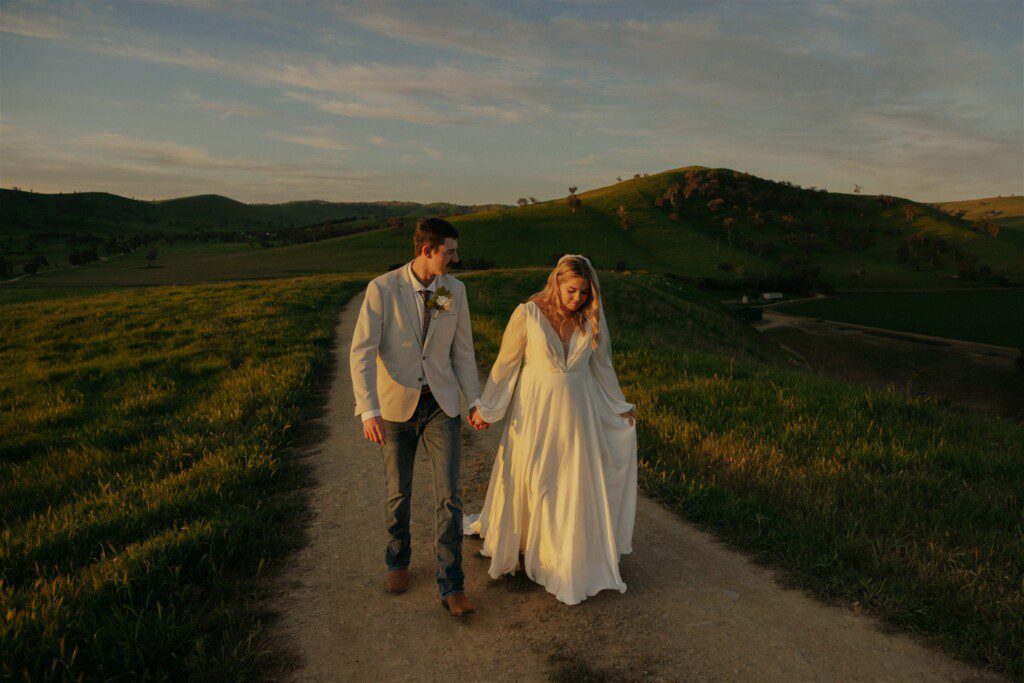 wedding couple holding hands walking up a dirt track in the country. Beautiful rolling hills in the background