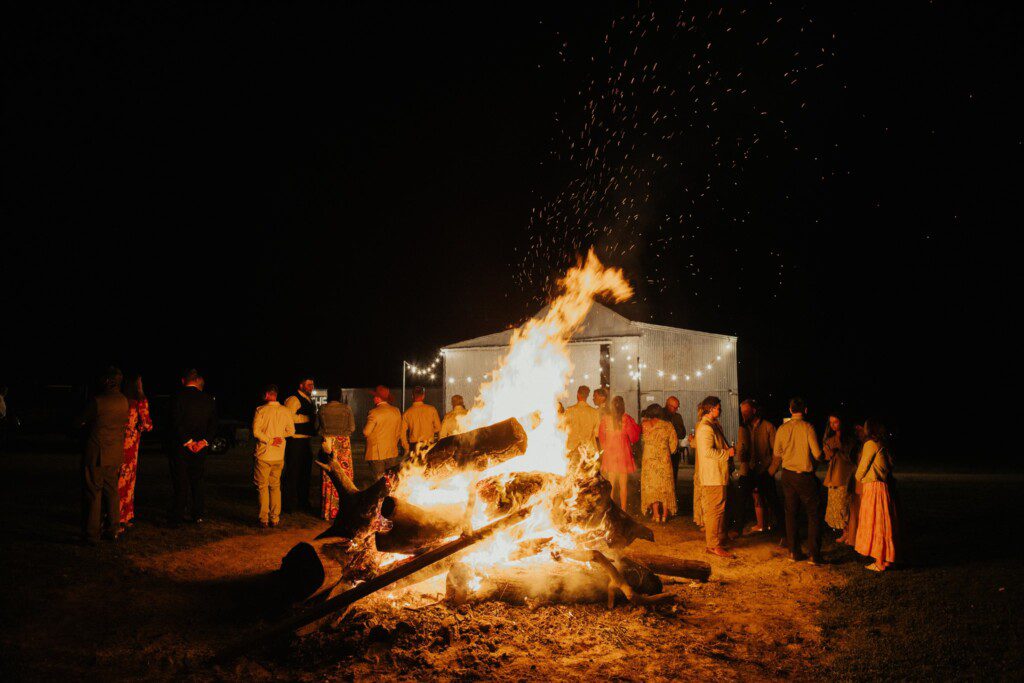 A huge bonfire roars with flame and wedding guests standing around its warmth. The grain shed in the background