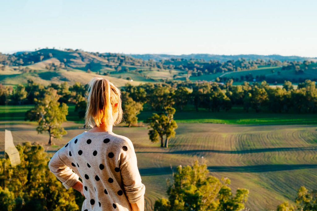 Woman standing with her back to the camera, hands on hips, overlooking a valley of rolling fields and trees.