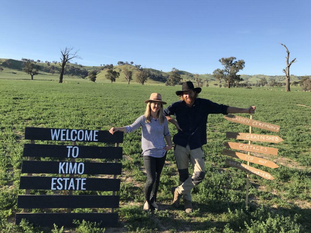 Two people with hats pose in a green field under a blue sky, beside a large 'Welcome to KIMO ESTATE' sign and wooden direction signs for nearby venues.