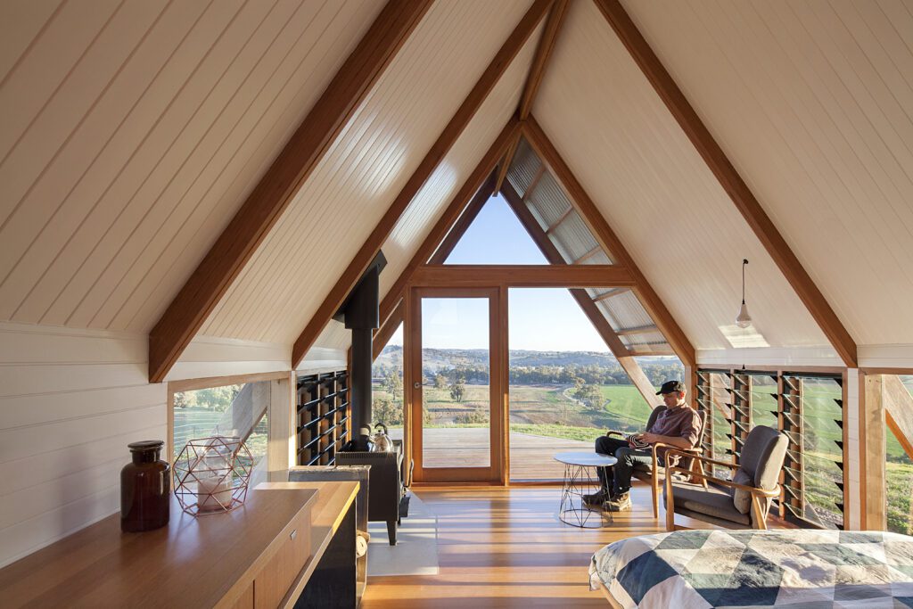 Loft-style room with exposed wooden beams and a large triangular window looking out on a hillside landscape; a man sits in a chair near the window.