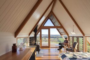 A-frame living space with tall triangular windows and exposed wooden beams; a man sits in a chair by the window looking outside, bright and airy.