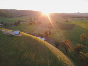 small a-frame off grid cabin on a hilltop from a drone. The sun is peeking over the horizon in the early morning. The best getaway from the city you can find in Australia